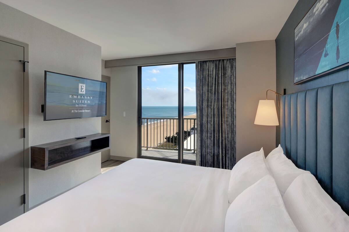 A hotel room with a large bed, a TV mounted on the wall, a balcony with a view of the beach, and a blue headboard.