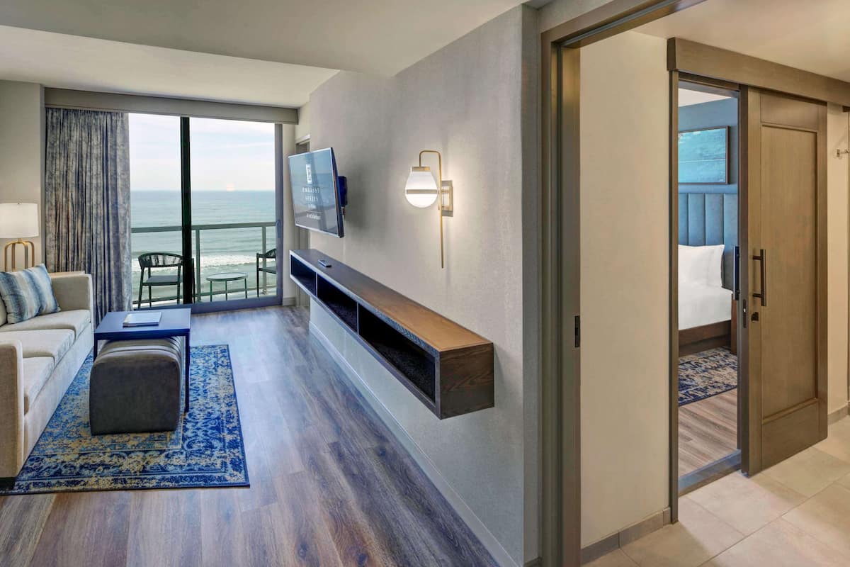 A modern hotel room with a balcony, a flat-screen TV, a wall-mounted light fixture, and a wooden bench.