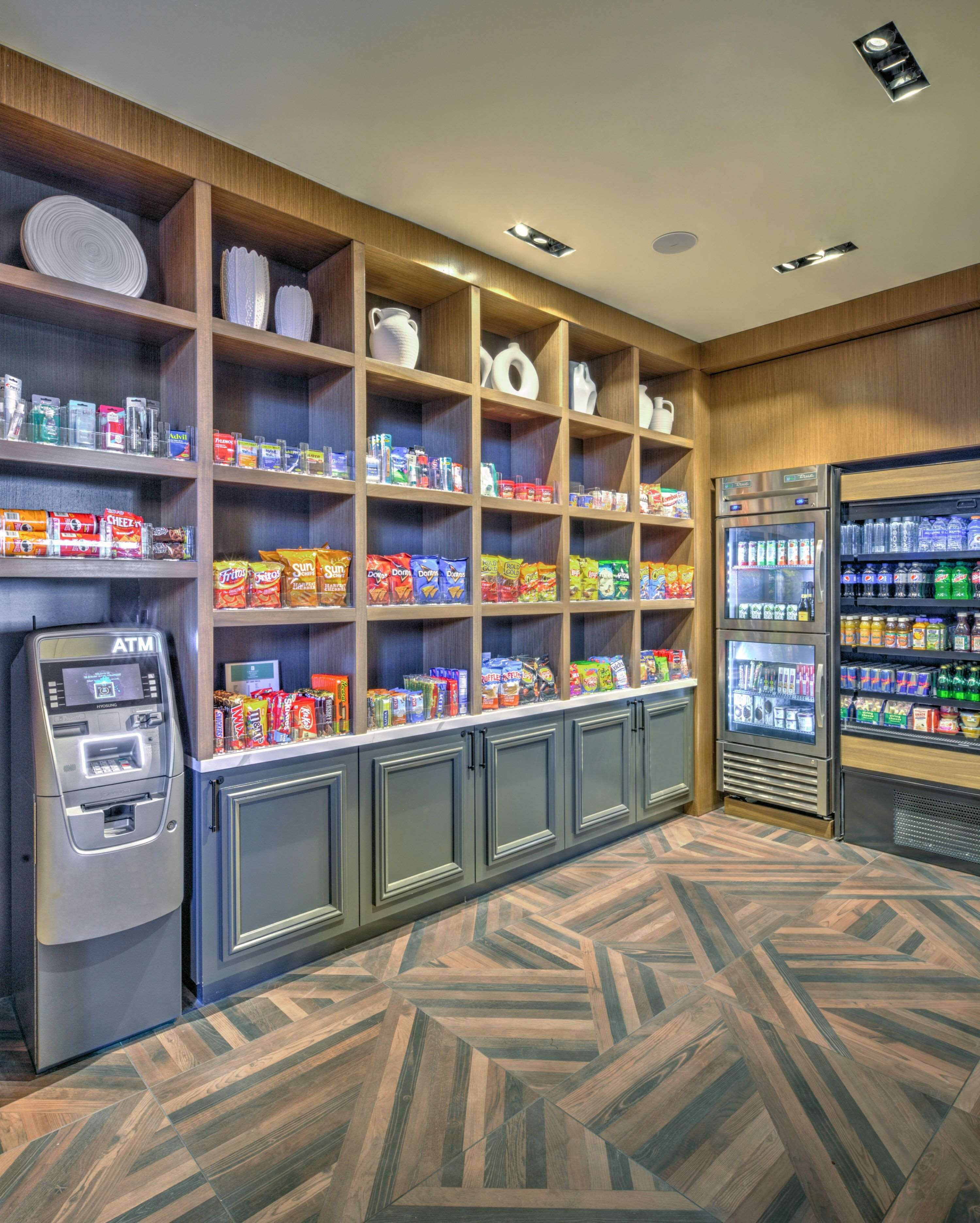 Et selvbetjeningsområde med en hæveautomat, hylder med snacks og drikkevarer, samt et mønstret gulv.
