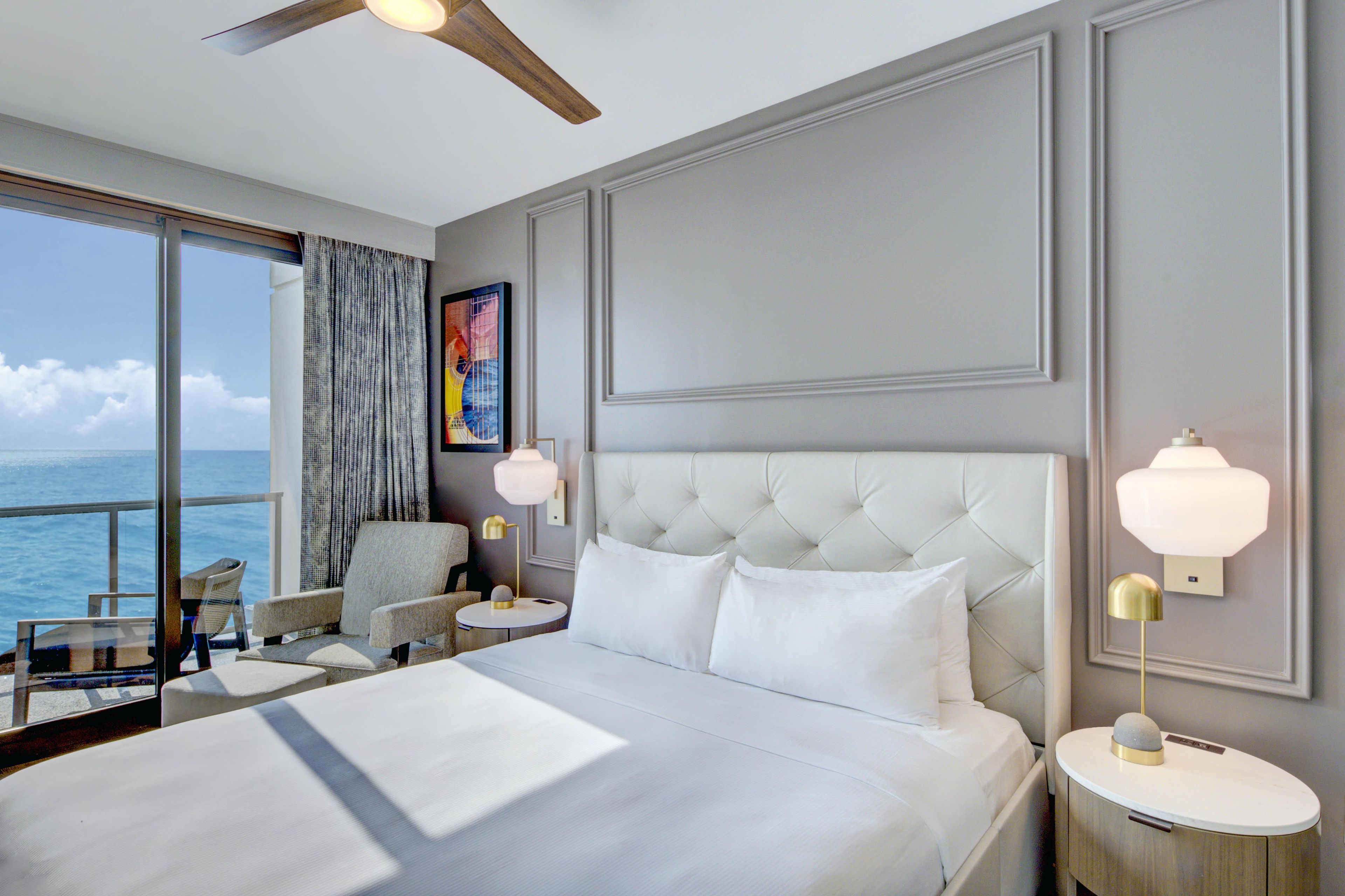 Ein Hotelzimmer mit einem großen Bett, Meerblick, Deckenventilator und einem Gemälde an der Wand.