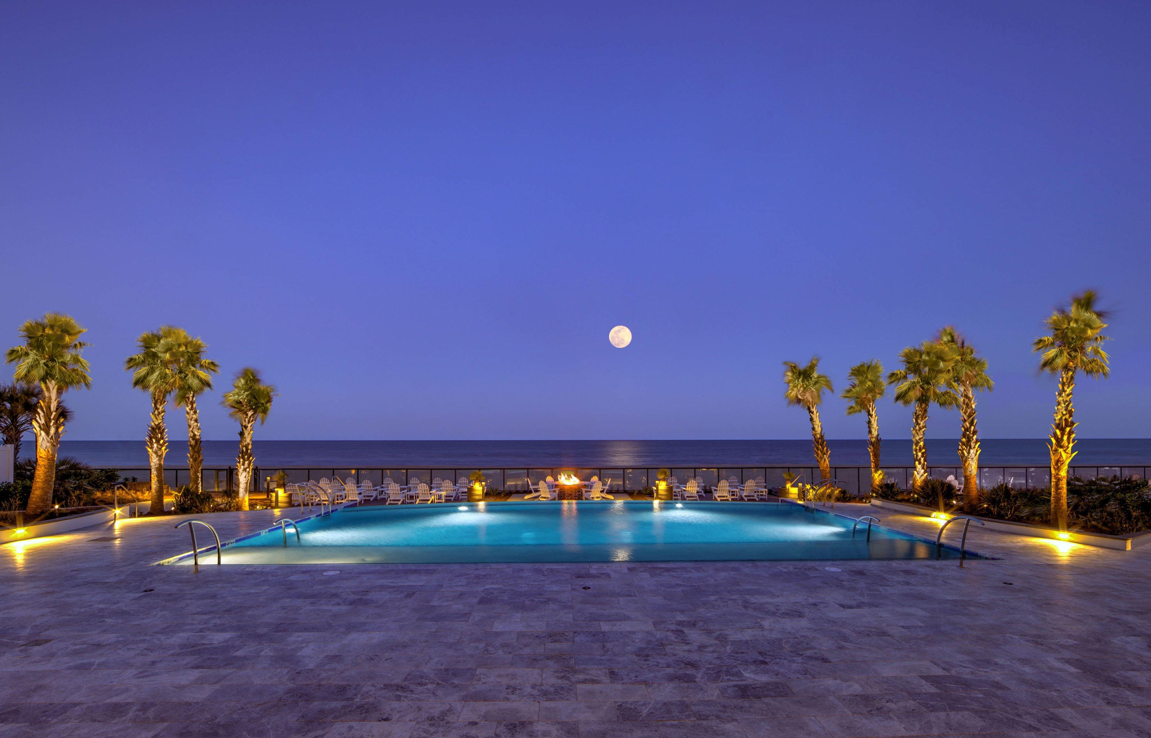 A poolside view with a moon in the sky, palm trees, and a view of the sea.