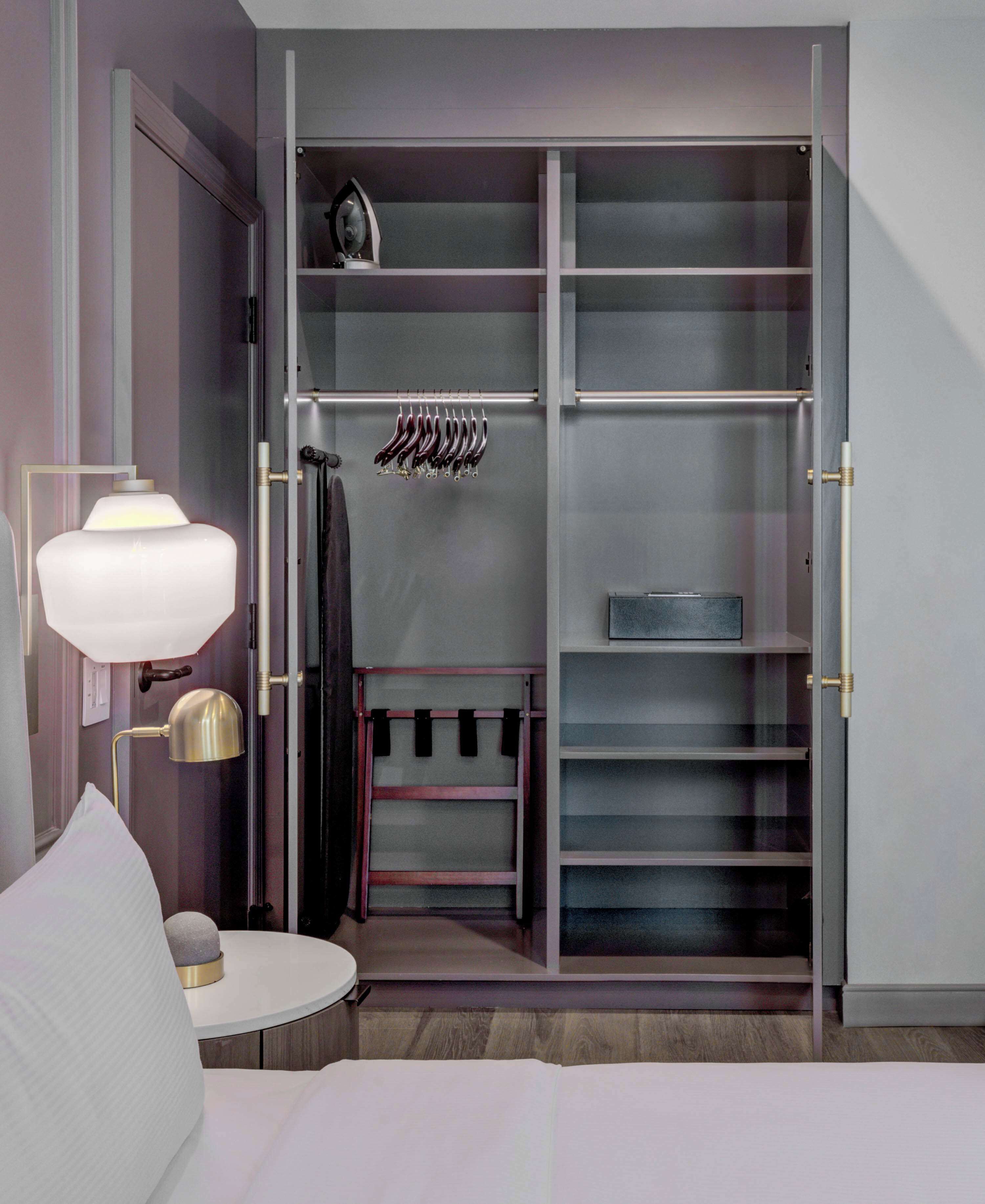 Un dormitorio con cama, mesita de noche, lámpara, armario con estantes y espacio para colgar, y una pequeña mesita auxiliar.