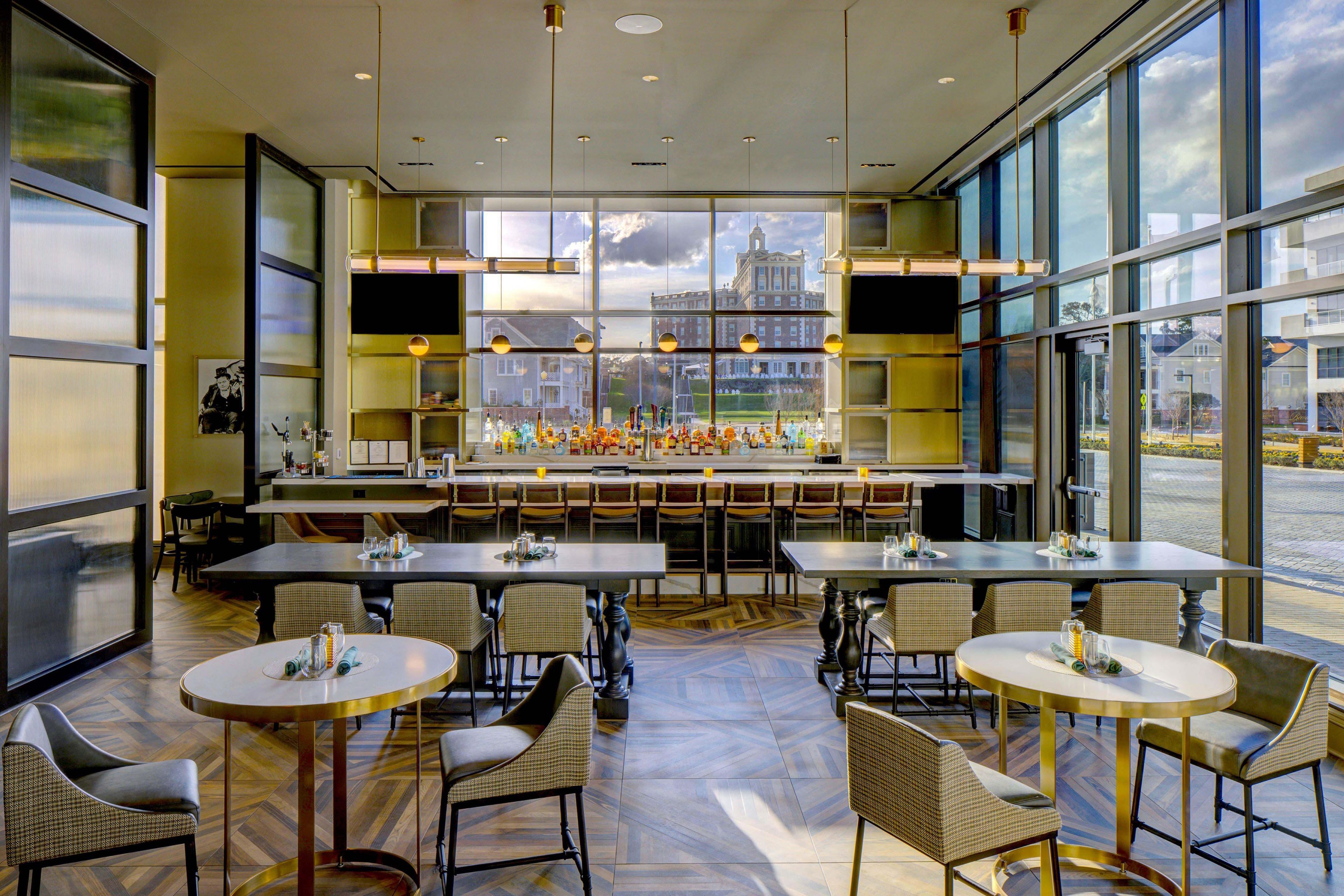 A restaurant with multiple tables and chairs, a bar area with bottles, and large windows offering a view of a building.