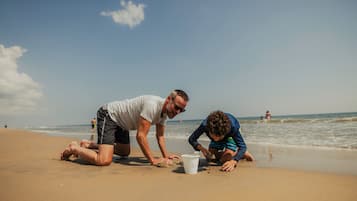 Two people on a sandy beach, one kneeling and the other standing, with a bucket between them.