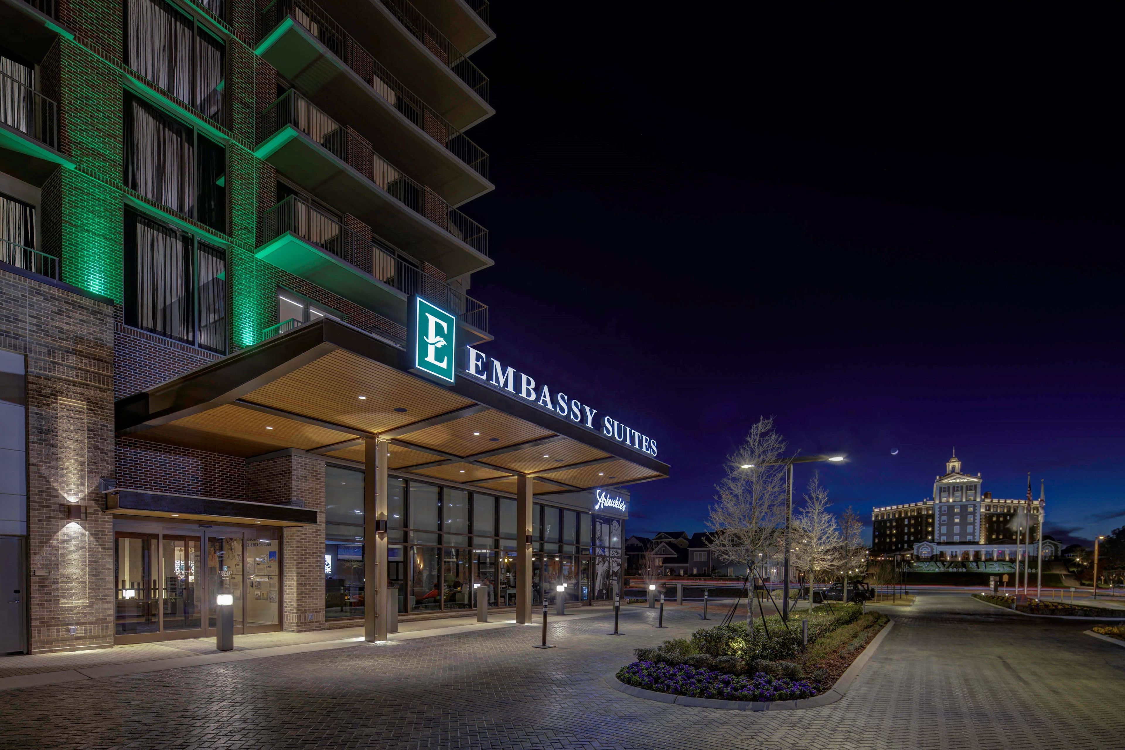 Entrada de un hotel con zona cubierta, un cartel que dice 'EMBASSY SUITES' y un edificio al fondo.