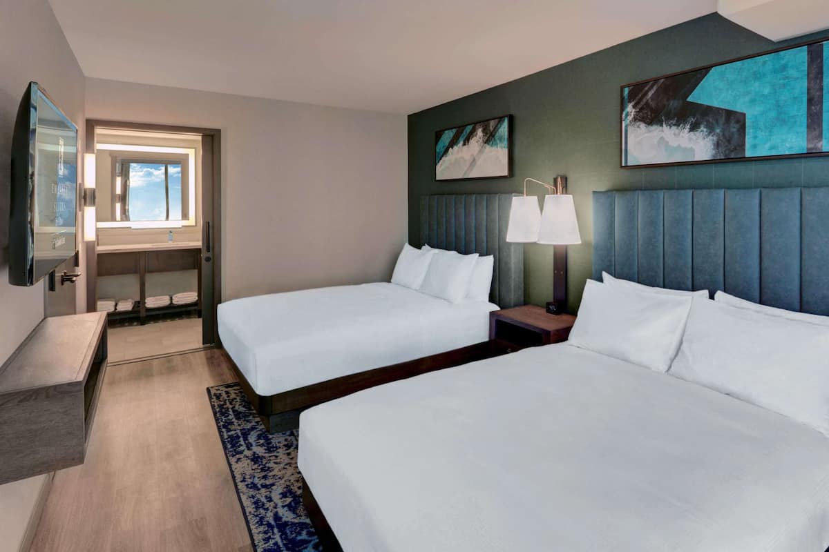 A hotel room with two beds, a nightstand with a lamp, a large window, and artwork on the walls.