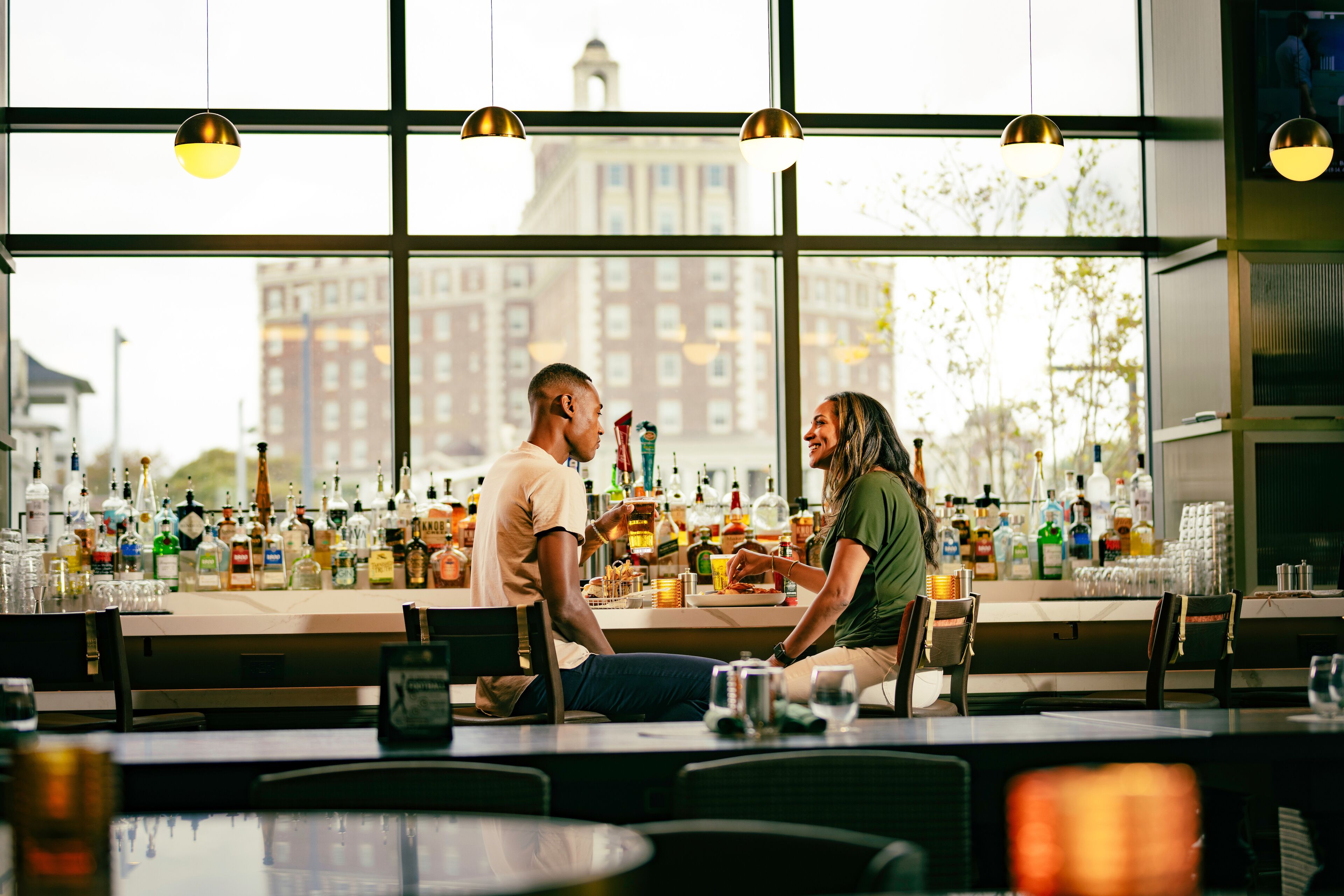 A bar with a view of buildings through large windows, bottles and glasses on the counter, and pendant lighting.