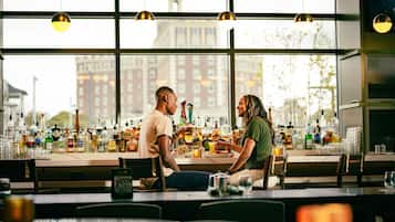 A bar with a view of buildings through large windows, bottles and glasses on the counter, and pendant lighting.