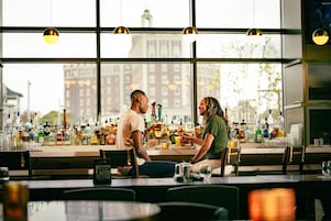 Um bar com vista para edifícios através de grandes janelas, garrafas e copos no balcão, e iluminação pendente.