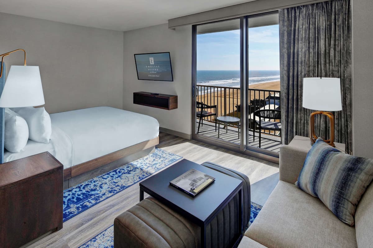 A hotel room with a bed, a sofa, a coffee table, a TV, and a balcony with a view of the beach.