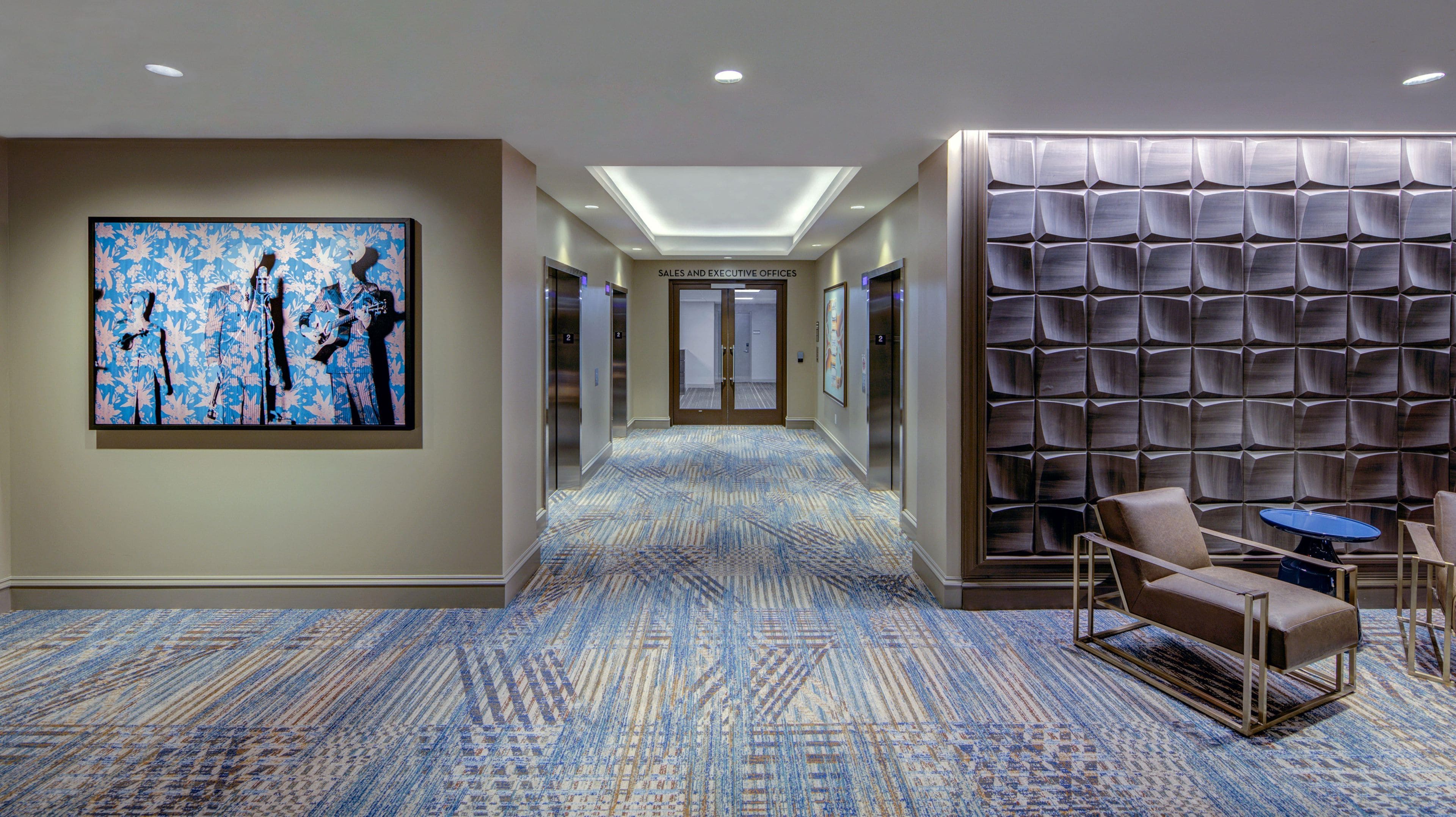 A hotel corridor with a patterned carpet, a large abstract painting, a seating area with a table, and a wall with a geometric design.