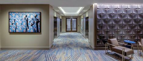 A hotel corridor with a patterned carpet, a large abstract painting, a seating area with a table, and a wall with a geometric design.