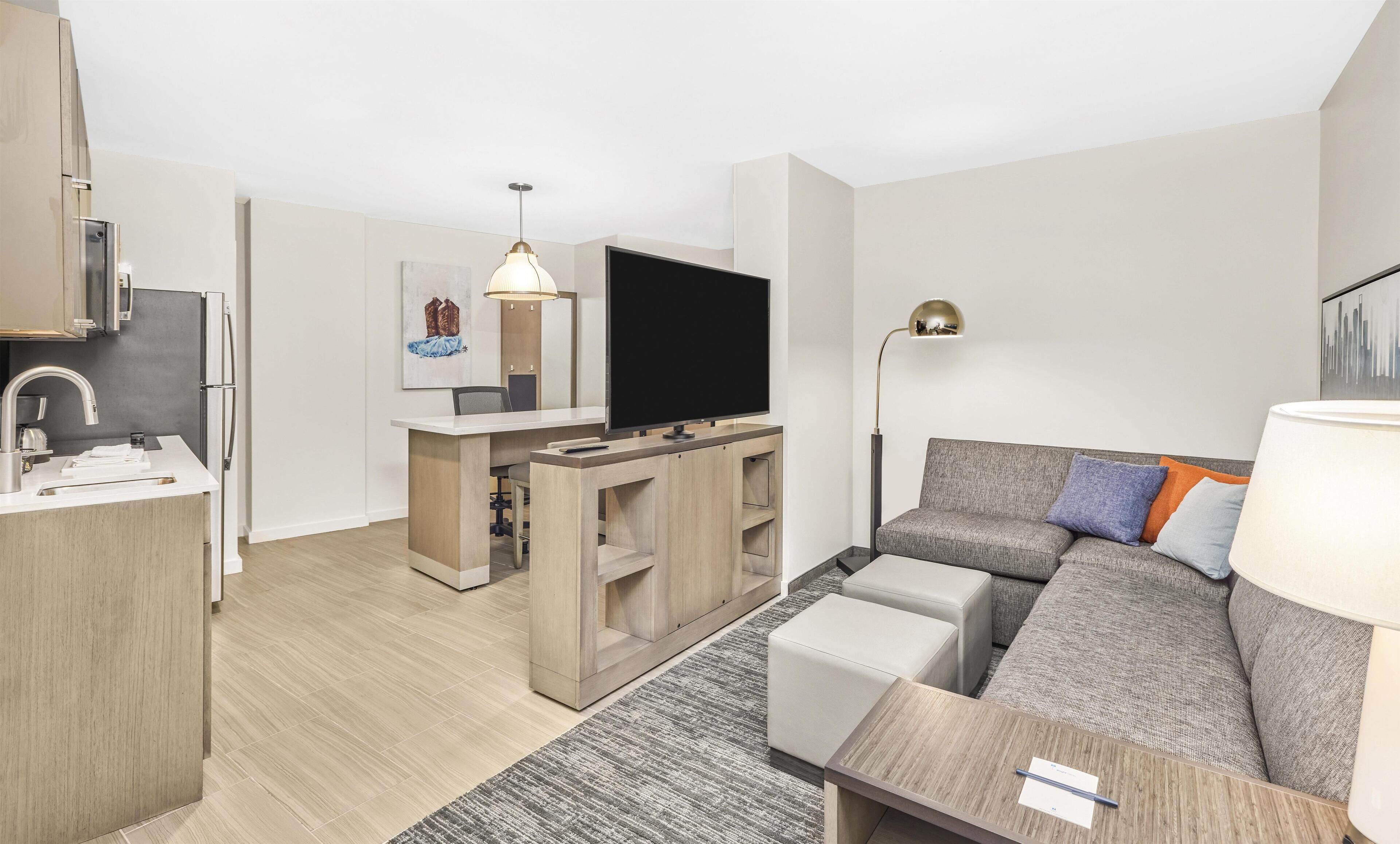 A modern living room with a sofa, a flat-screen TV, a dining area with a table and chairs, and a kitchen with a refrigerator and sink.