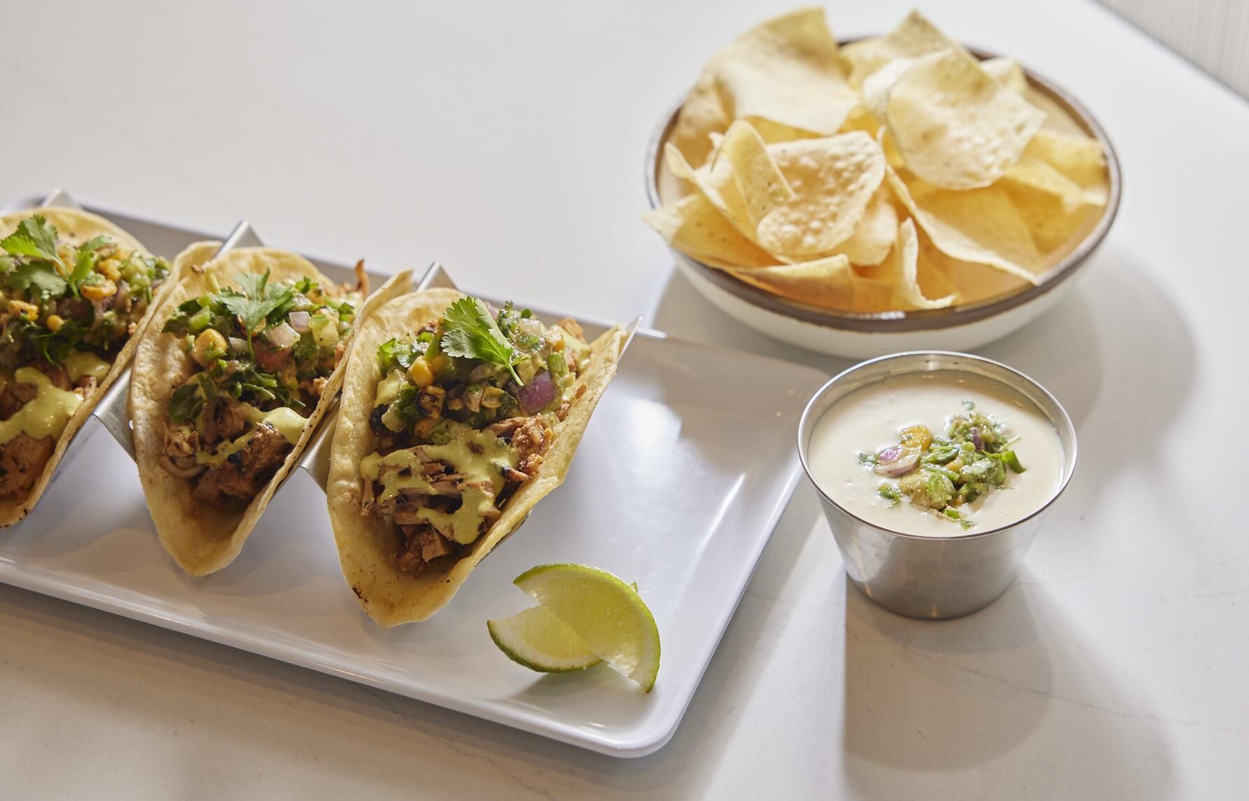 Three tacos with toppings and a bowl of chips on a white tray.
