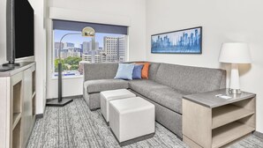 Habitación de hotel moderna con sofá seccional gris, televisor de pantalla plana, escritorio con lámpara y vista panorámica de la ciudad.