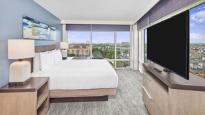 Habitación de hotel con una cama grande, un televisor de pantalla plana, un cuadro en la pared y vistas a la ciudad a través de amplios ventanales.