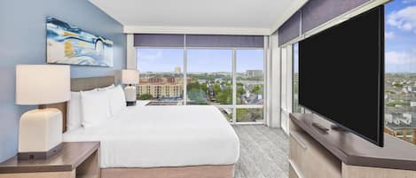 Ein Hotelzimmer mit einem großen Bett, einem Flachbildfernseher, einem Gemälde an der Wand und Blick auf die Stadt durch große Fenster.
