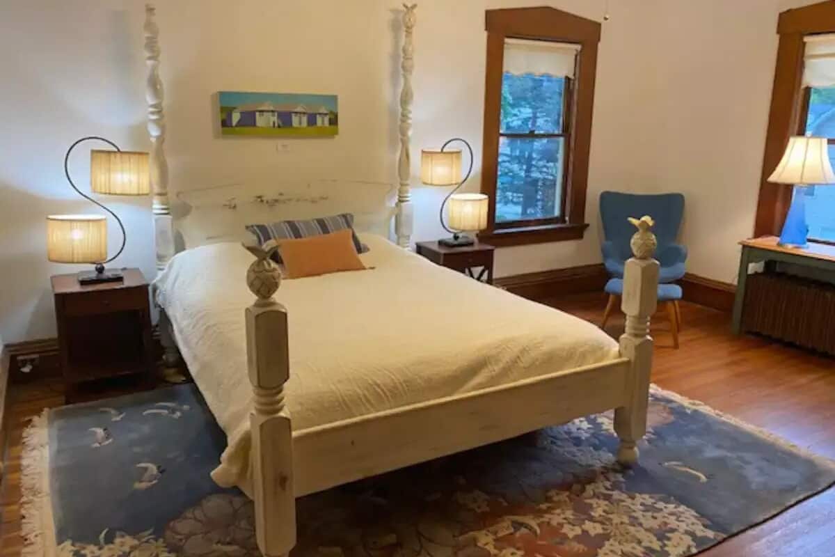 A bedroom with a four-poster bed, two bedside lamps, a window with a view, and a chair.