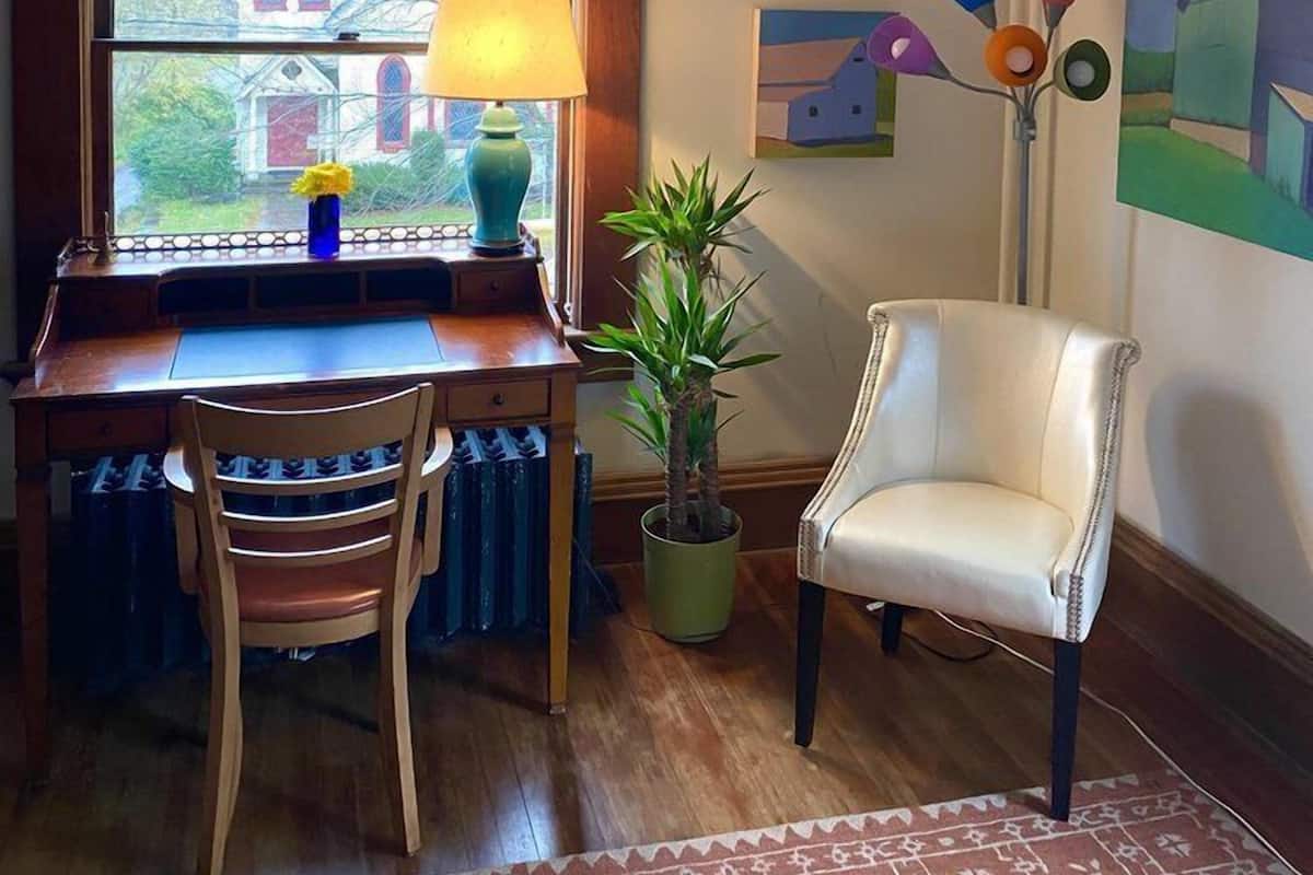A cozy room with a wooden desk, a chair, a window with a view of a house, a lamp, a plant, and a painting on the wall.