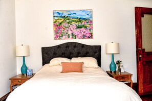 A neatly made bed with a headboard, two bedside tables with lamps, a painting on the wall, and a wooden door.