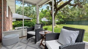 A covered patio with wicker furniture, a small table with a plant, and a view of a green backyard. - Beautiful Backyard! | Angel Oak in Park Circle (North Charleston)