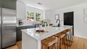 A modern kitchen with a large island, bar stools, stainless steel appliances, and a window with a view of greenery. - Beautiful Backyard! | Angel Oak in Park Circle (North Charleston)