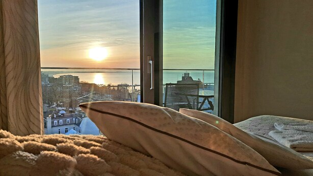 Ein Schlafzimmer mit großem Fenster, das Blick auf eine Stadtlandschaft und ein Gewässer bei Sonnenuntergang bietet.