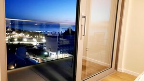 Blick vom Balkon mit Glastür auf eine Skyline mit beleuchteten Gebäuden und einem Gewässer.