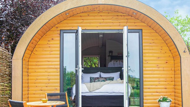 Een knus houten huisje met een open deur die uitkomt op een bed met kussens en een dekbed. Daarbuiten staat een kleine tafel met stoelen en een bloempot.