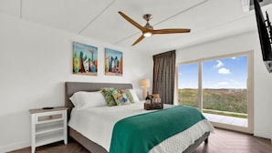 Ein Schlafzimmer mit Bett, Nachttisch, Deckenventilator, Fenster mit Blick und Kunstwerken an der Wand.