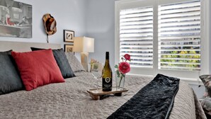 Ein Schlafzimmer mit einem Bett, einem Nachttisch mit einer Weinflasche, einer Vase mit Blumen und einem Fenster mit Jalousien.