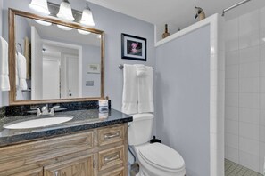 A bathroom with a granite countertop sink, wooden vanity, white toilet, and a shower area. - Relaxed Island Escape with Pool and Hot Tub Access (South Padre Island)