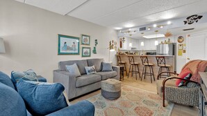 Ein modernes Wohnzimmer mit grauem Sofa, blauen Sesseln und einem gemusterten Teppich. Im Hintergrund ist ein Küchenbereich mit Barhocker und einem Esstisch zu sehen.