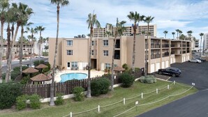 Ein mehrstöckiges Hotel mit Pool, umgeben von Palmen und einem eingezäunten Parkplatz.