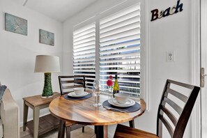 A dining area with a round wooden table, two chairs, a lamp, and a view through white blinds. - Beach Steps Away: Garden Oasis with Pool & Jacuzzi (South Padre Island)