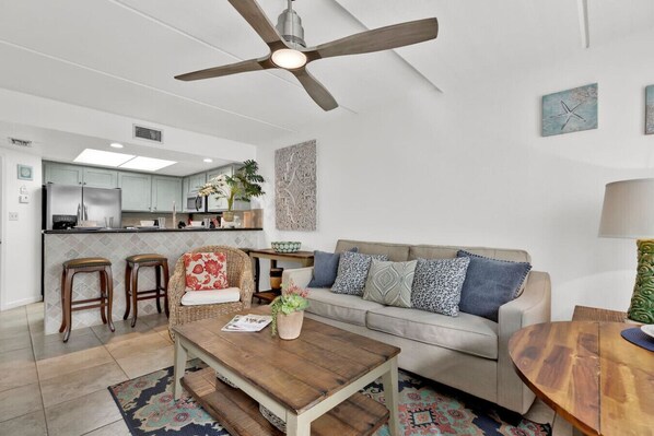 A modern living room with a sofa, coffee table, and two barstools. A ceiling fan and kitchen are visible in the background.