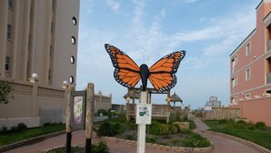 Una escultura de mariposa en un área ajardinada con senderos y edificios al fondo.