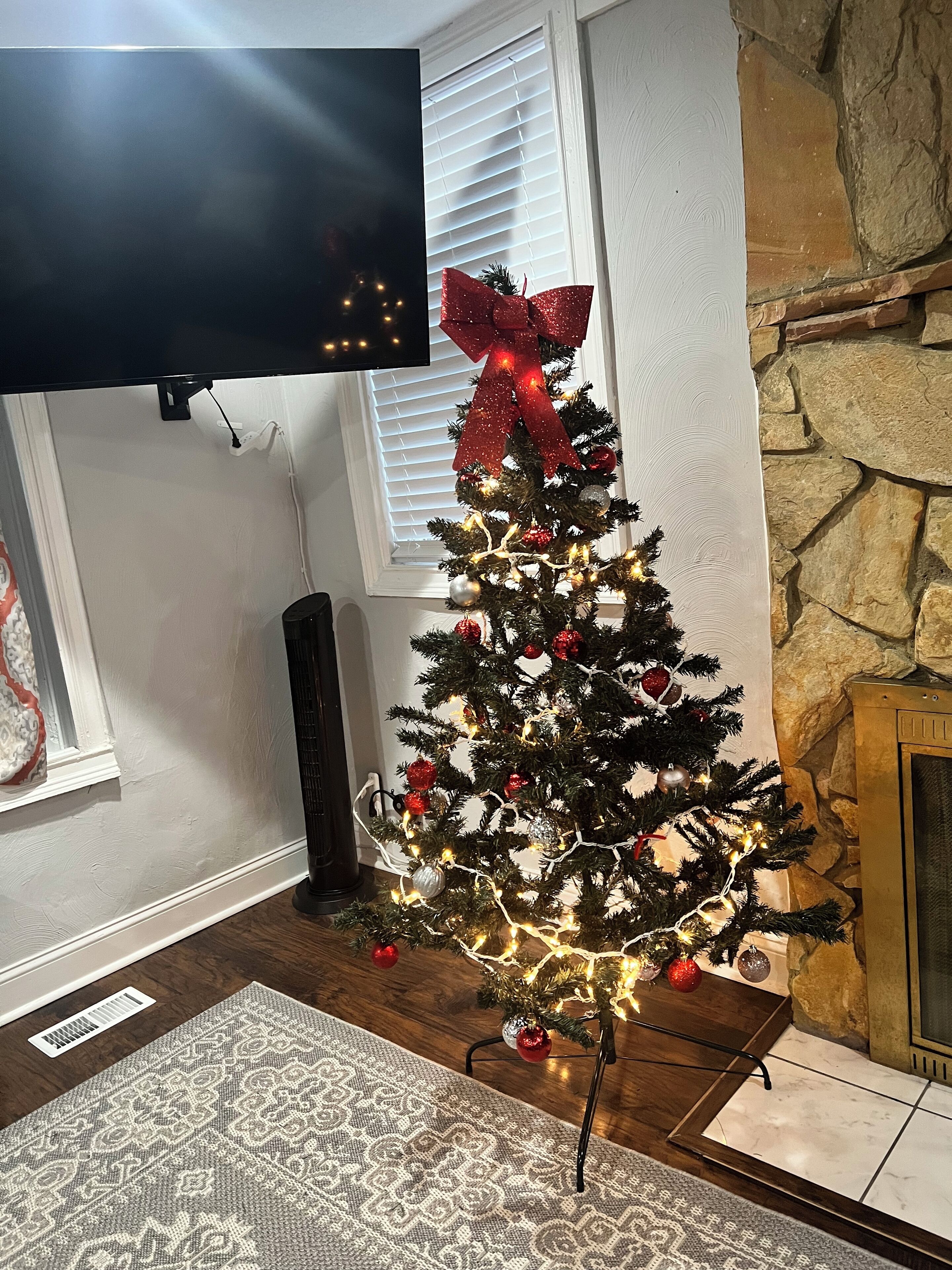 Un árbol de Navidad con luces y adornos, ubicado en una sala de estar con chimenea de piedra, un televisor montado arriba y una alfombra estampada en el suelo.