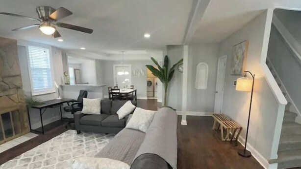 Ein geräumiges Wohnzimmer mit dunklem Holzboden, einem grauen Ecksofa, einem Couchtisch mit Pflanze und einem Deckenventilator.