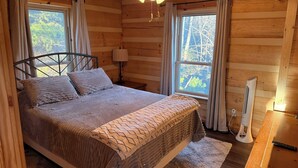 A wooden cabin bedroom with a bed, a ceiling fan, two windows, and a heater.
