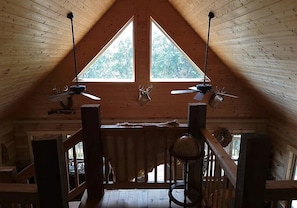 Une pièce mansardée en bois avec deux fenêtres, un ventilateur de plafond et une table.