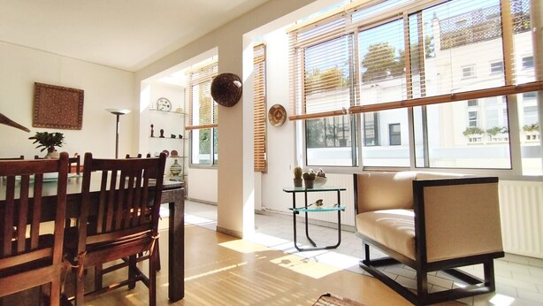 A spacious living area with a dining table, chairs, a sofa, and large windows providing natural light.