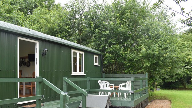 A green building with a door open, revealing a room with a table and chairs. There are steps leading to a raised platform with more chairs.