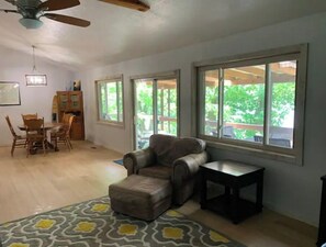A spacious living room with a large window, a ceiling fan, a dining area with a table and chairs, and a patterned rug.