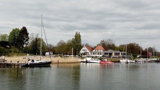 A lakeside view with boats docked, a building with a red roof, and trees in the background.
