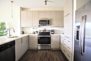 A modern kitchen with stainless steel appliances, light wood cabinetry, and a dark wood floor.