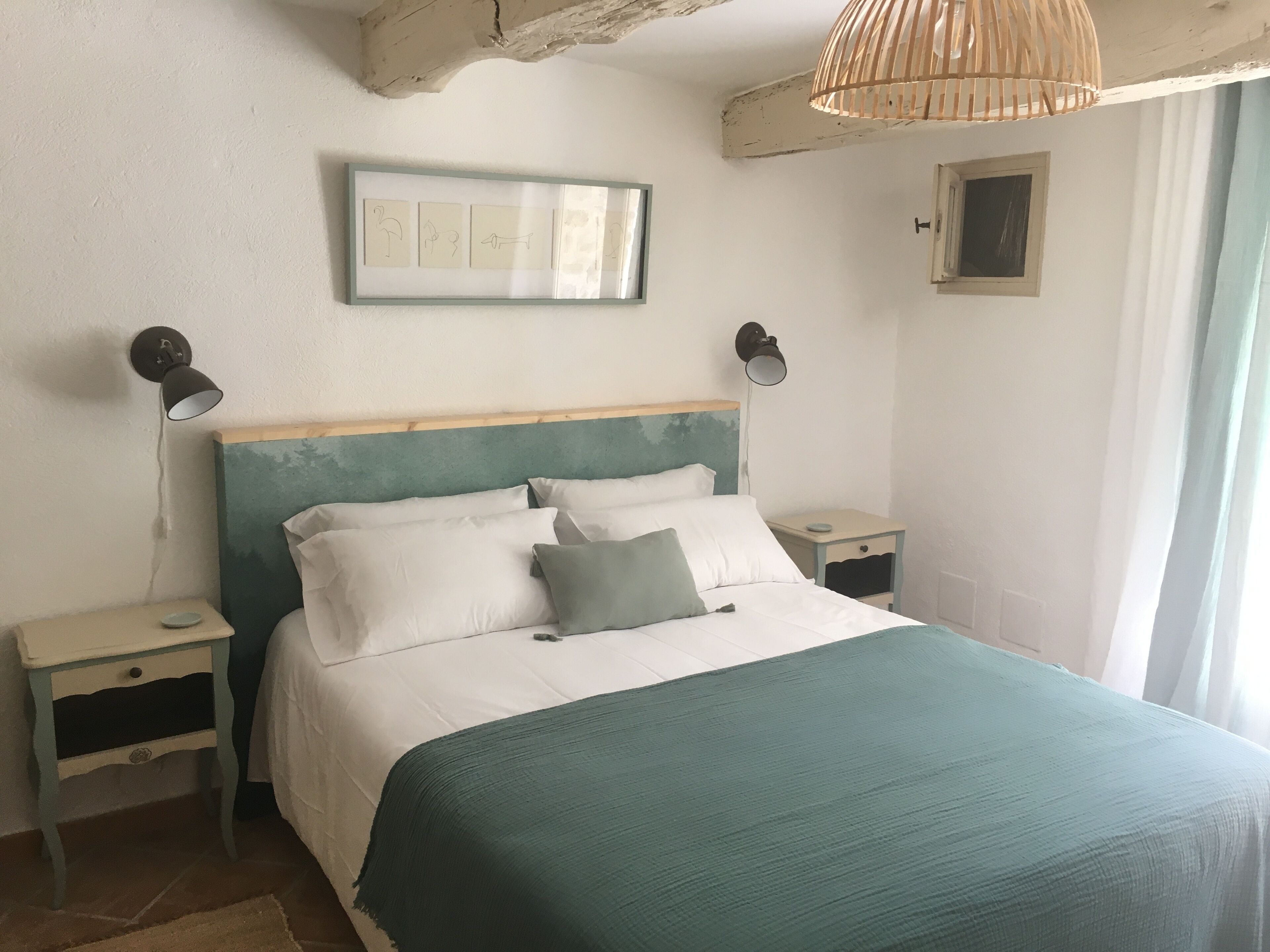 A bedroom with a bed, two bedside tables, a wall-mounted light fixture, a framed picture, and a window with curtains.