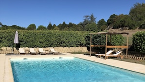 En rektangulär swimmingpool med klart, blått vatten, omgiven av beige kakelplattor. Det finns vita solstolar och en pergola med ett tak av trä.