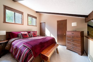 Une chambre avec un grand lit, une commode en bois et un banc. Il y a deux fenêtres et une porte.