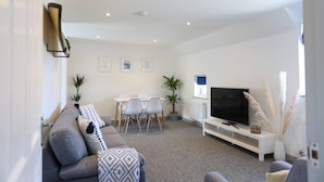 A modern living room with a grey sofa, a dining table with chairs, a flat-screen TV, and decorative plants. - Shorebreak - Bracklesham Bay Apartment (Chichester)